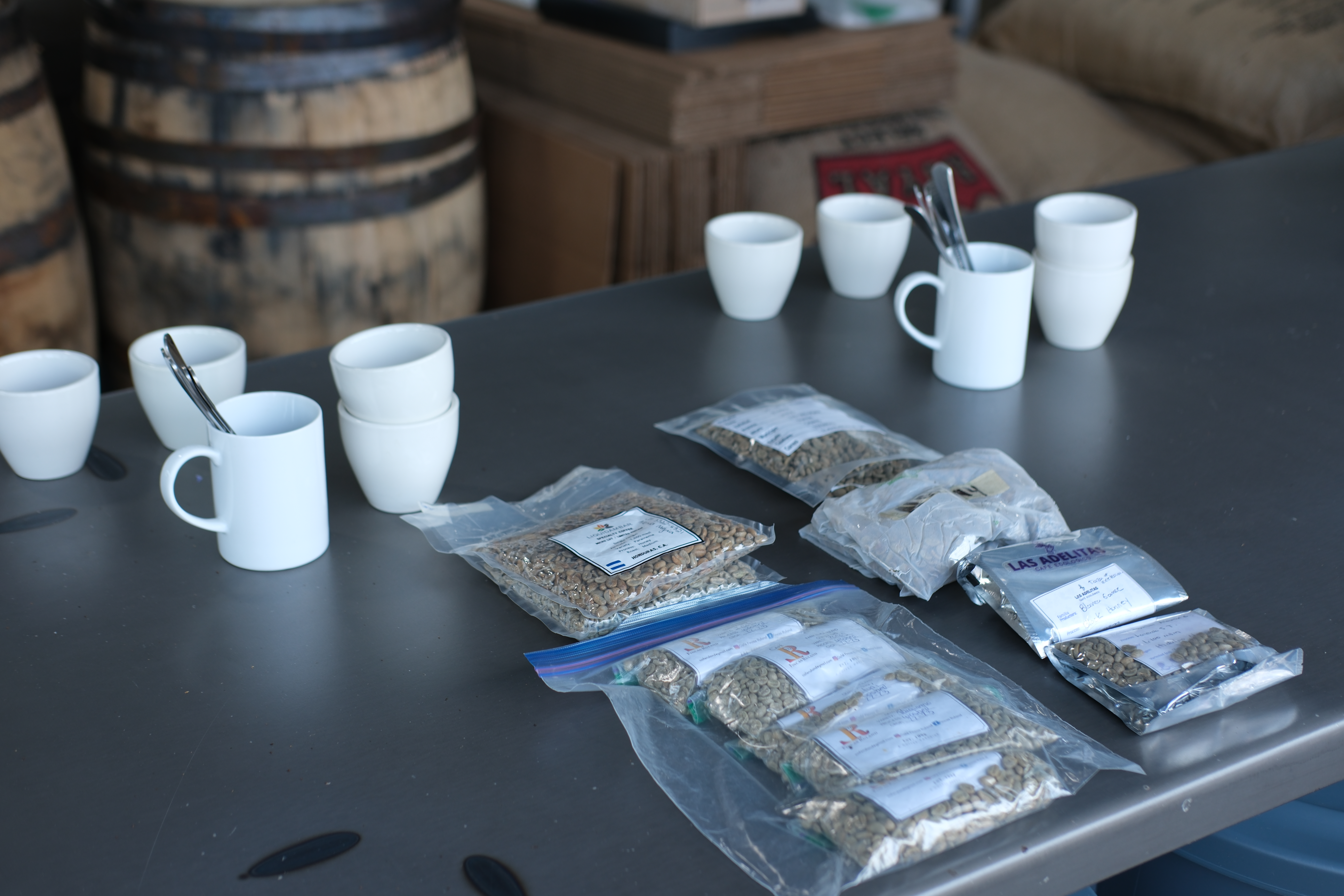 Freezer bags of coffee and cupping cups and spoons on a table with barrels of coffee in the background