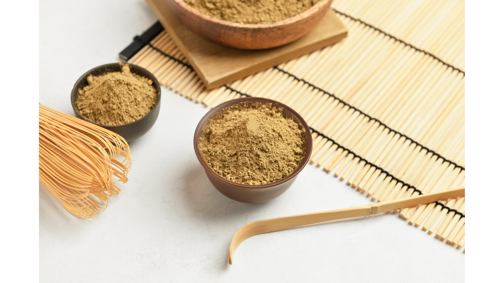 Three bowls of matcha powder, a bamboo whisk, and a matcha scoop on a bamboo mat