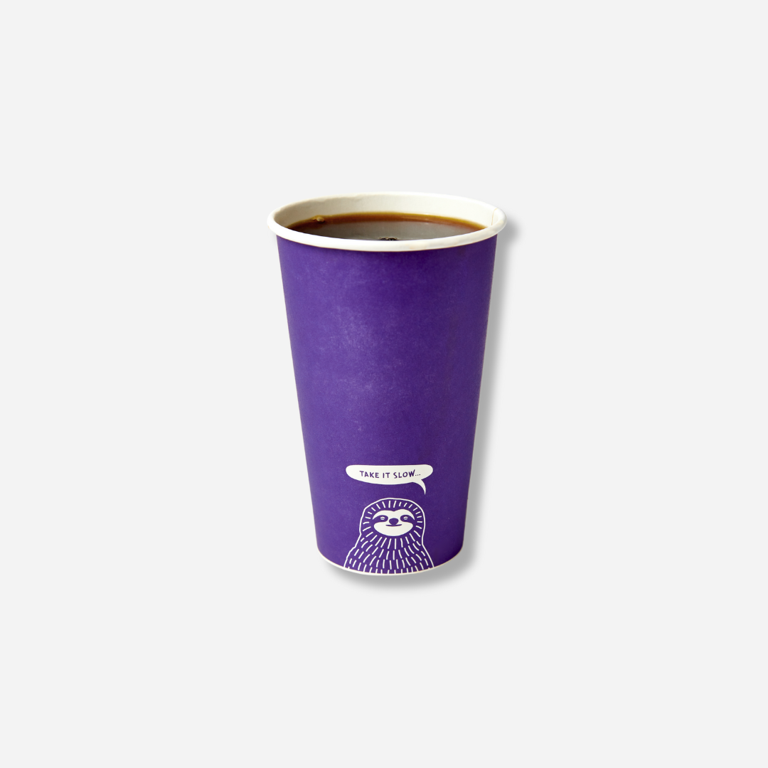 A tall purple paper cup filled with hot black coffee, with a small white sloth graphic near the bottom and a speech bubble that says “TAKE IT SLOW…”