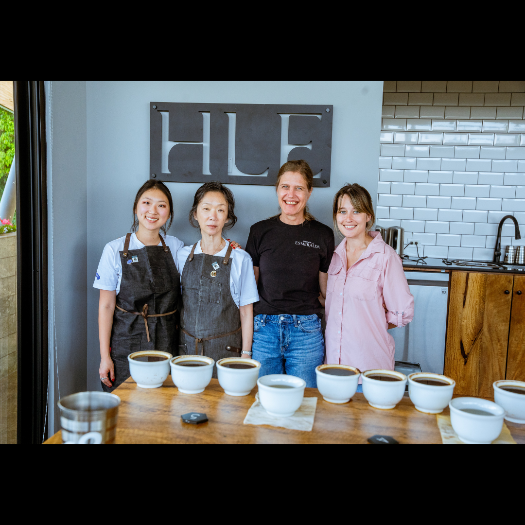 Four people stand smiling behind a cupping table with several bowls of coffee in front of them, inside a bright coffee lab with a sign reading “HLE” on the wall.