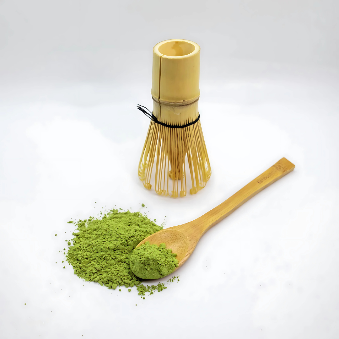 A bamboo matcha whisk standing upright next to a wooden spoon filled with green matcha powder.