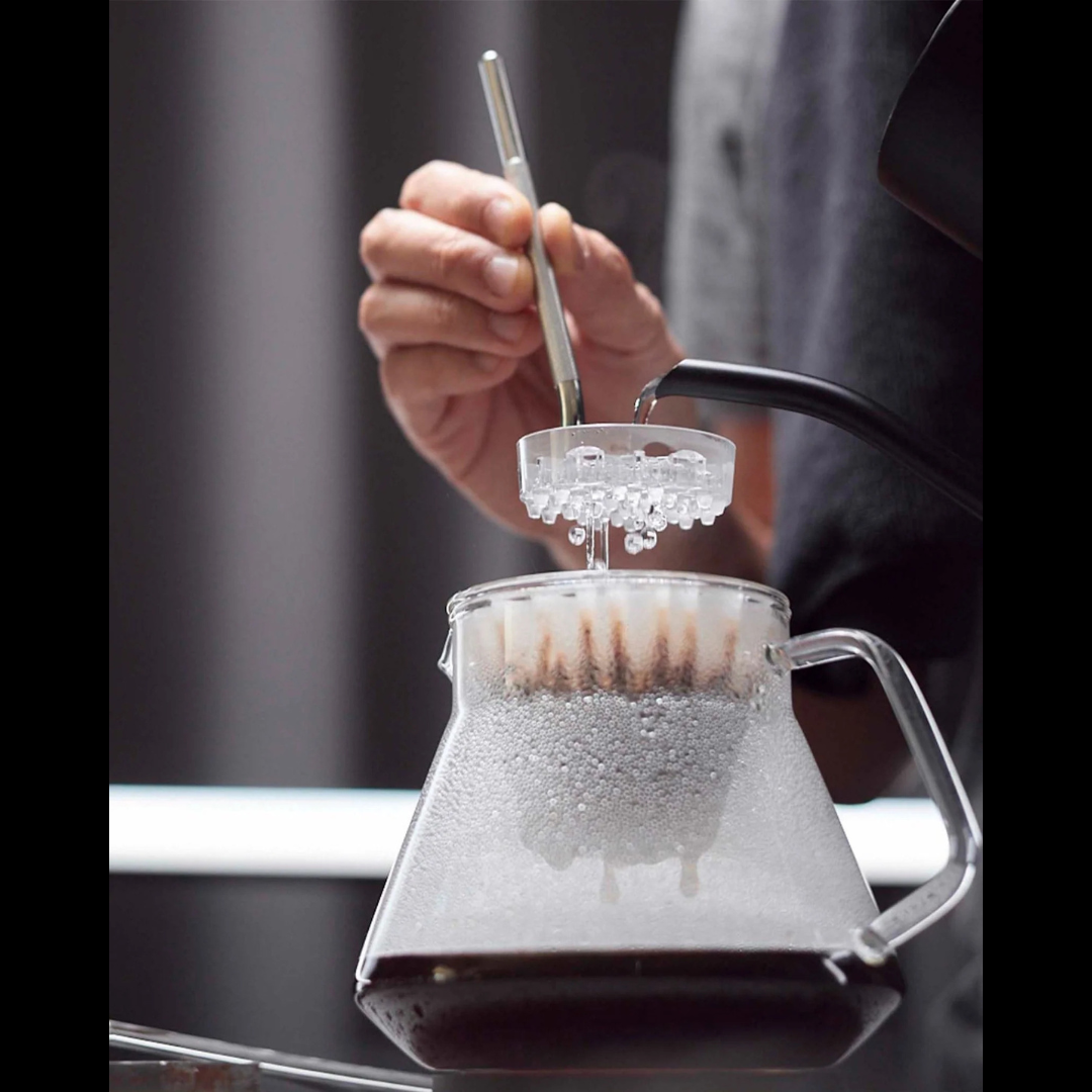 A person pours hot water from a kettle over a Melodrip tool positioned above a glass pour over dripper, allowing the water to fall gently onto the coffee grounds.