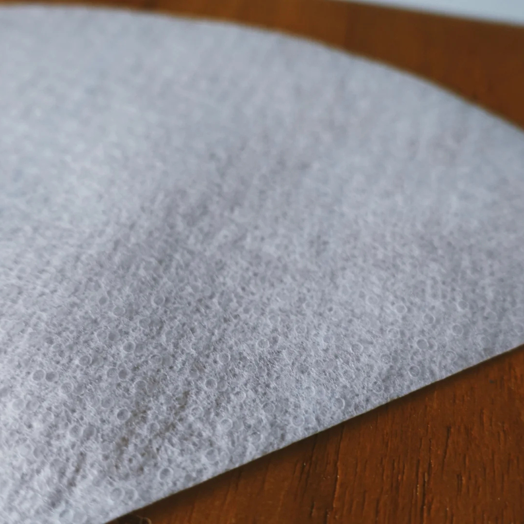 A close-up image of a white, V-shaped non-woven coffee filter. The filter’s surface shows a textured pattern of tiny interlocked circles, highlighting its unique porous structure designed for efficient water flow and clarity in brewing.